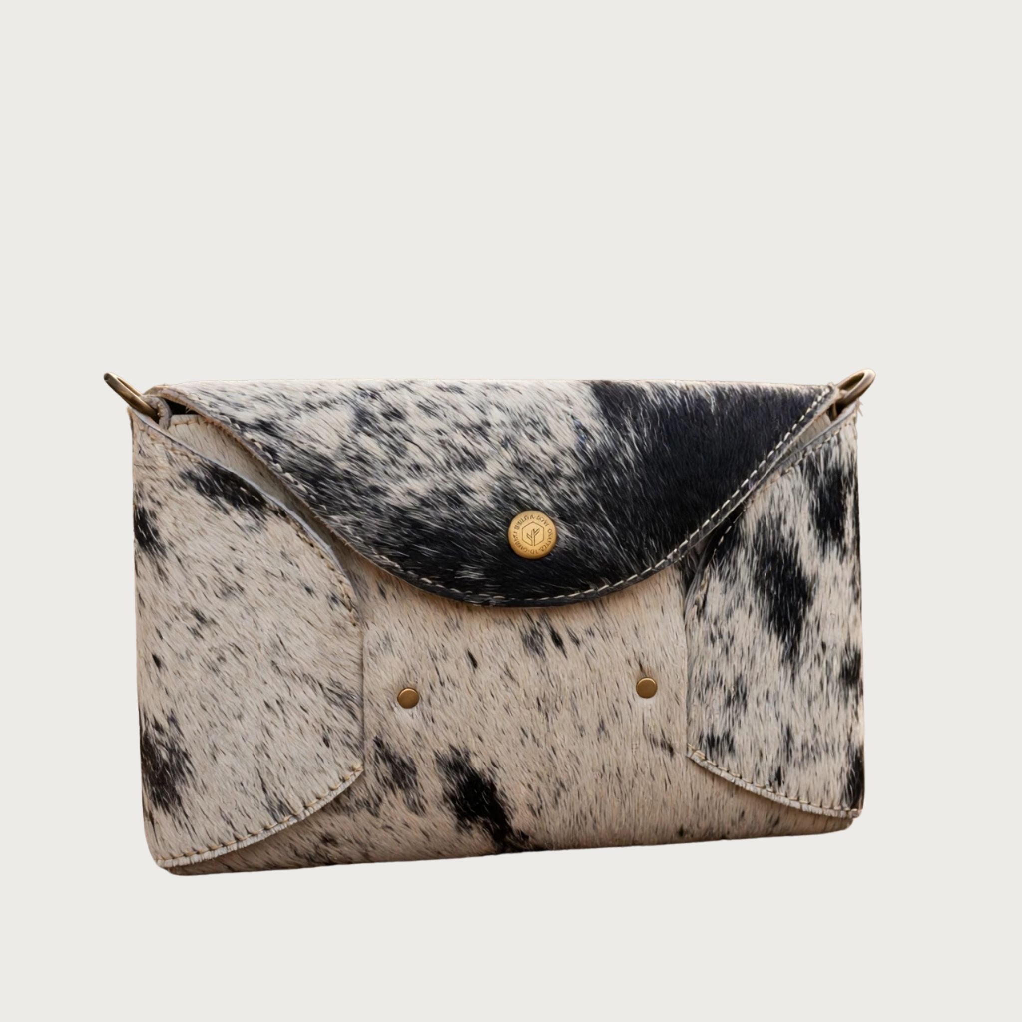 Three-quarter angle view of Oak & Elk hair-on-hide cowhide crossbody bag on white background, showing bag depth and antique brass hardware