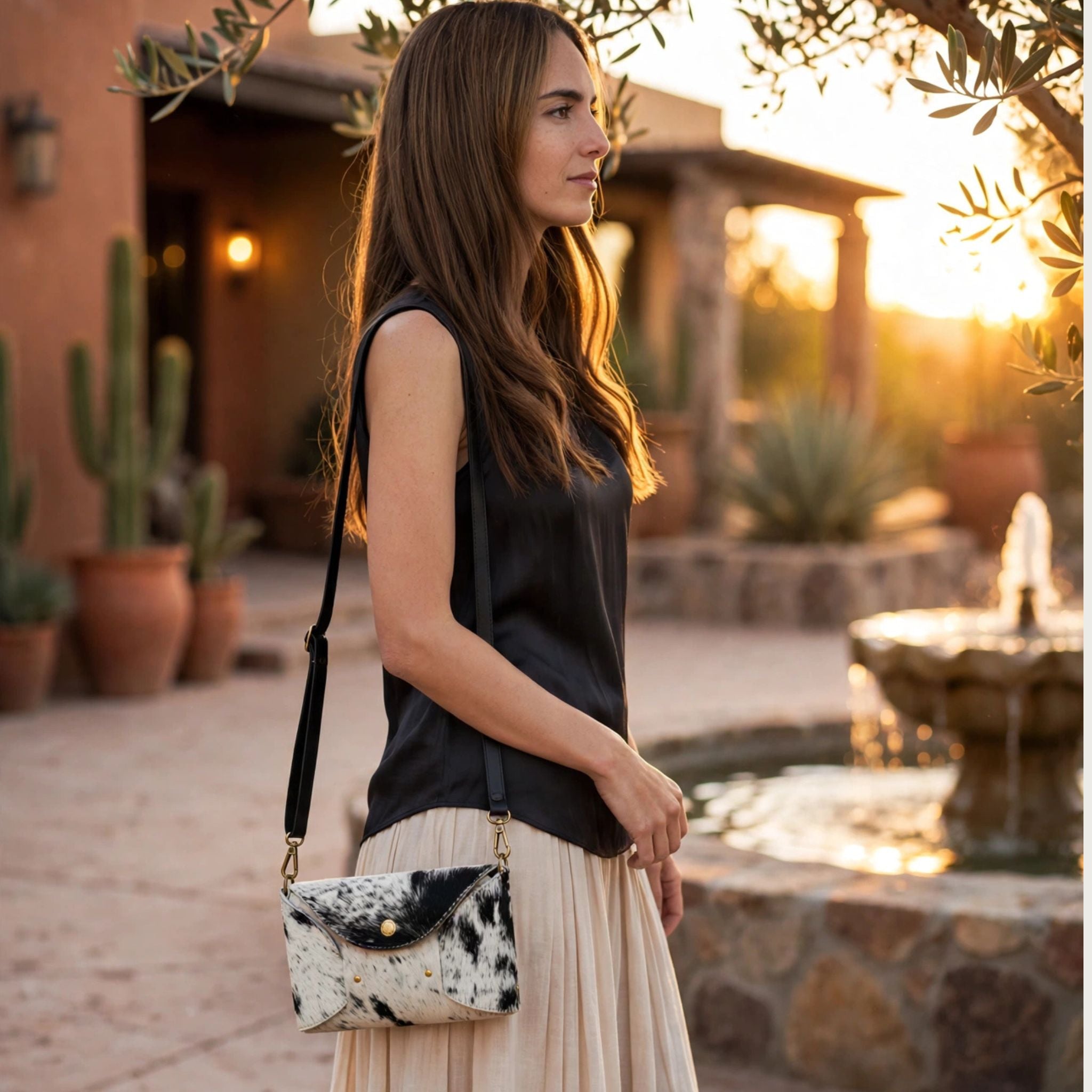 Woman wearing Oak & Elk PALOMA cowhide crossbody bag with black leather strap, black sleeveless top and beige pleated midi skirt, profile view in desert hacienda courtyard at sunset