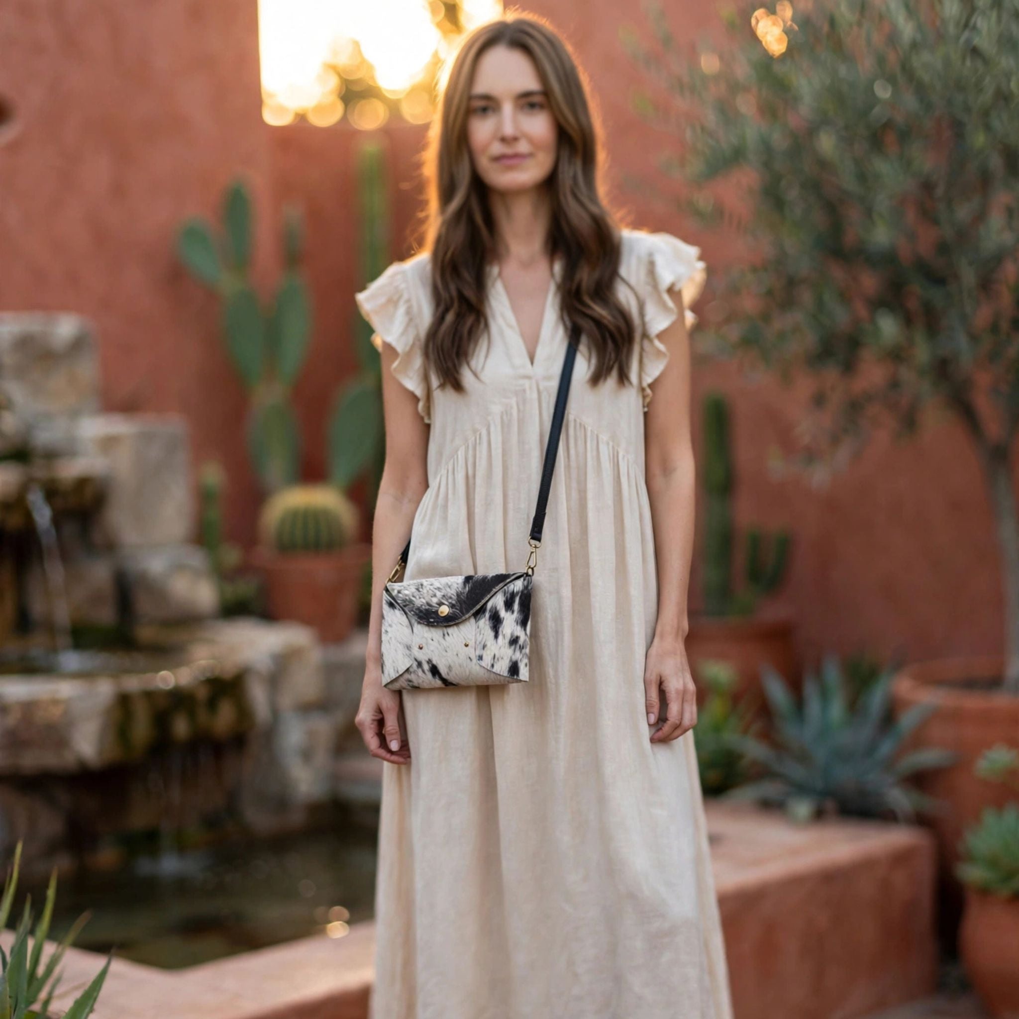 Woman in beige ruffle flutter-sleeve dress wearing Oak & Elk PALOMA handmade cowhide crossbody bag with black leather strap, frontal view in desert hacienda courtyard at golden hour
